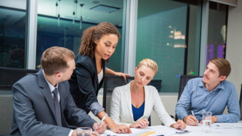 African American businesswoman standing in meeting room, pointing to document and explaining business results. Business team are looking at her and listening with attention. Working concept