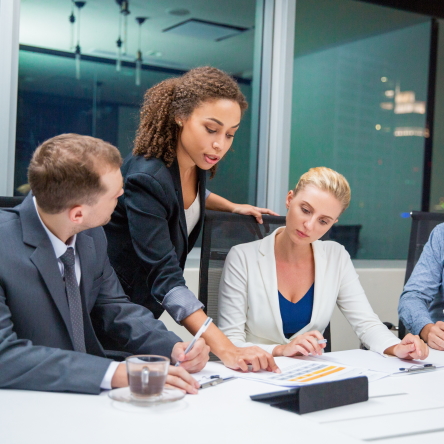 African American businesswoman standing in meeting room, pointing to document and explaining business results. Business team are looking at her and listening with attention. Working concept