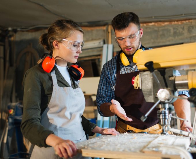 Waist-up portrait of attractive apprentice standing at drill press and listening to her male mentor with attention, he explaining her operating principle of machine